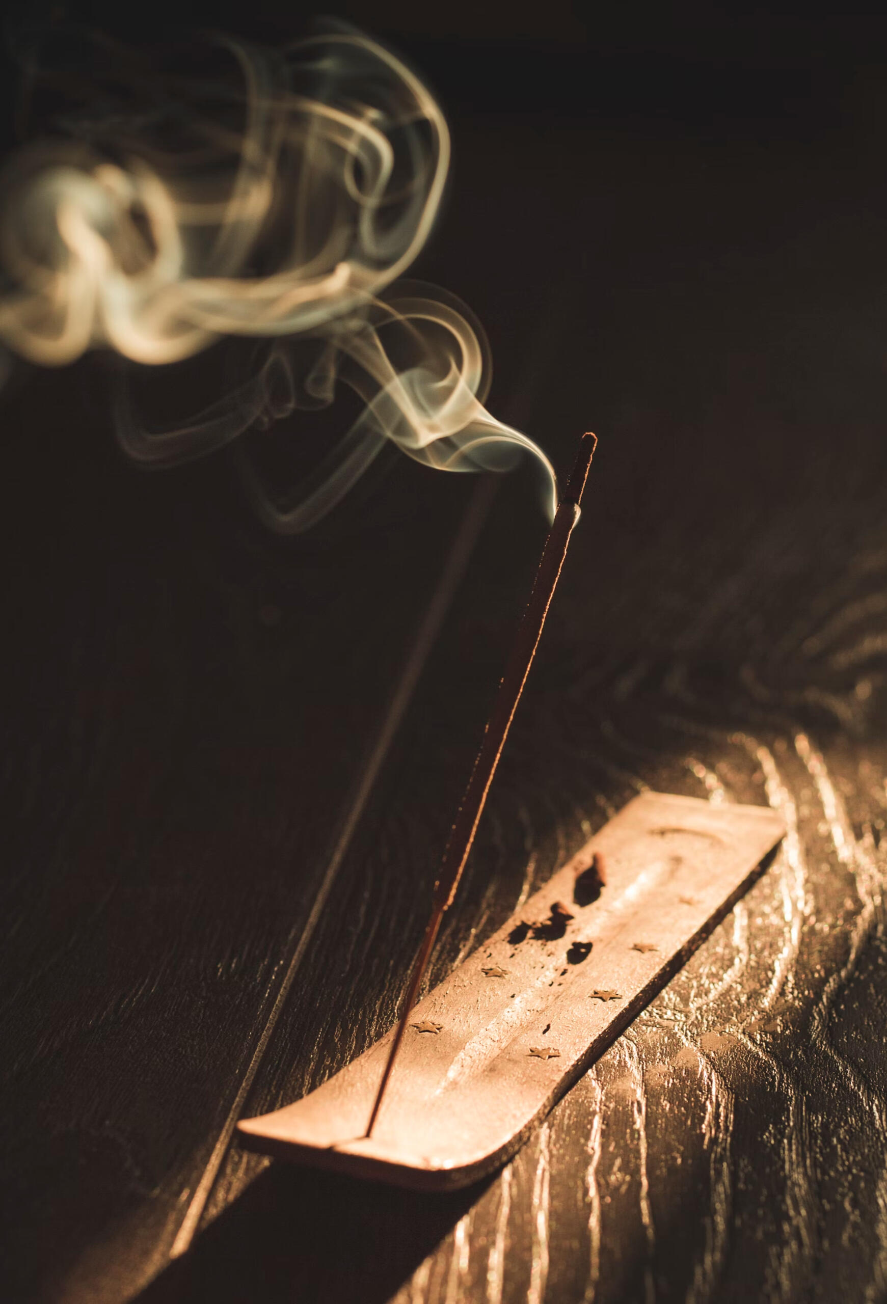A single stick of incense in a wooden incense burner on a wooden floor, caught in a beam of sunlight, smoke swirling in the air.